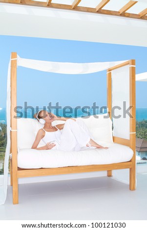 Woman reclining on a white canopied seat in the summer sunshine on the patio enjoying music on her headphones with an ocean backdrop