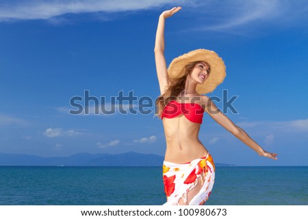 Beautiful lively woman full of the joys of life twirling with her arms in the air in a colourful bikini and sarong against an ocean backdrop