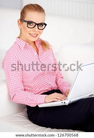 Pretty blond girl sitting on sofa with laptop and wearing black glasses.