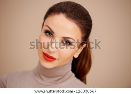 Gorgeous beautiful brunette with her hair tied back in a ponytail wearing a poloneck and red lipstick  studio headshot on a beige background