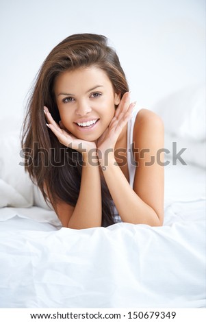 Vivacious young woman in bed lying on the bedclothes on her stomach with her chin on her hands giving the camera a beautiful smile