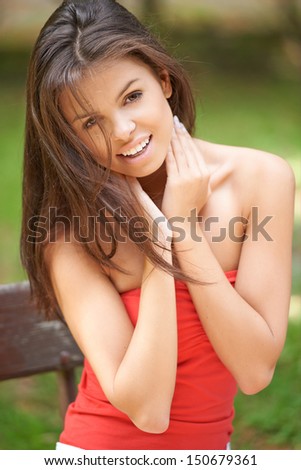 Happy attractive young brunette woman in a red summer top sitting on a bench outdoors in the garden