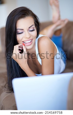 Beautiful young woman smiling in amusement as she reads the screen of her laptop while relaxing on a sofa at home