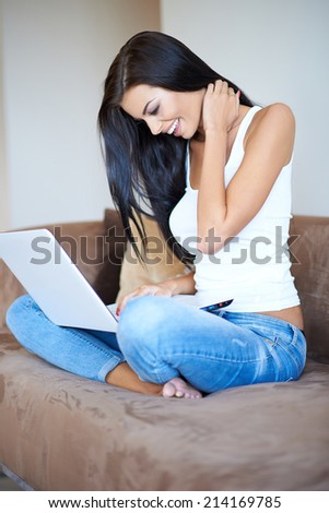 Casual sexy young woman sitting cross legged on a sofa smiling happily as she browses the internet and social media on a laptop
