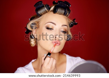 Beautiful young woman applying makeup with her blond hair in curlers as she carefully applies lipstick while looking in a mirror