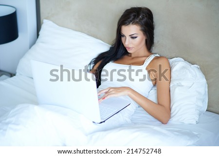 Beautiful woman working in bed lying typing on a laptop computer as she relaxes against the white pillows  high angle view