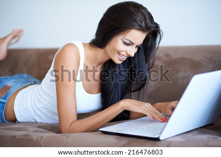 Young woman lying on her stomach a sofa using a laptop computer to surf the internet with a happy relaxed smile