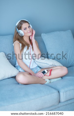 Happy young woman relaxing listening to music sitting cross-legged on a sofa at home with her MP3 storage device in her hand
