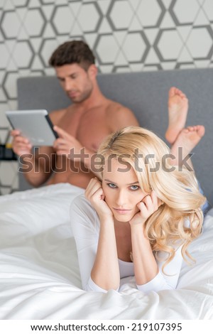 Disgruntled woman lying on her bed glaring at the camera because her husband is ignoring her to read his tablet computer