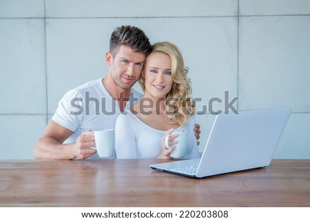 Attractive couple having a mug of coffee together sitting at a wooden table with a laptop computer looking at the camera with a smile