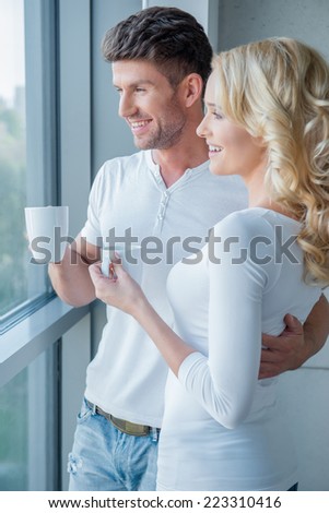 Young couple standing enjoying the view through a large glass window as they enjoy an early morning mug of coffee