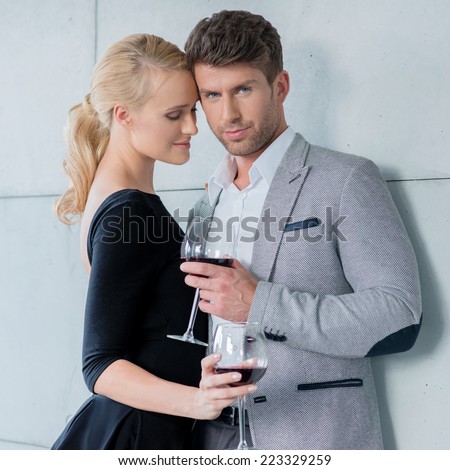 Elegant couple on a romantic evening out standing arm in arm drinking red wine with the handsome young man looking at the camera