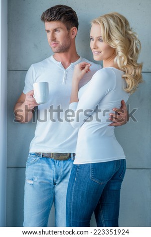 Attractive couple standing close together in an affectionate embrace looking out of a window as the man holds a mug of coffee