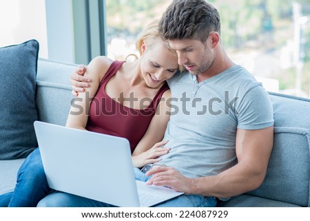 Loving couple surfing the web on a laptop computer as they sit arm in arm on the sofa at home
