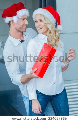 Playful man giving his wife a Christmas gift surprising her as he reaches round from behind in their colorful red Santa hats