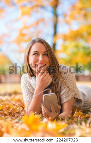Pretty Smiling Lady Lying on Dry Leaves at Grassy Ground with Mobile Phone. Looking at Right Side Frame.