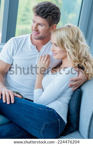 Happy young couple relaxing on a couch at home smiling as they sit in a close embrace watching something off the left of the frame