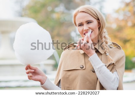 Pretty blond woman eating candy floss biting on a sticky morsel with a smile as she stands outdoors in an autumn park