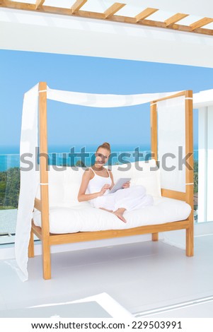 Elegant happy woman sitting on a canopied day bed on an outdoor tropical patio smiling at the camera