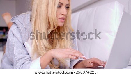 Young Blond Woman in Gray Sleep Robe Lying on her Stomach on White Couch While Surfing the Internet Using her Laptop Computer.