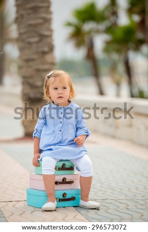 Cute little girl sitting on a stack of colorful small suitcases on a sidewalk with tropical palm trees looking at the camera