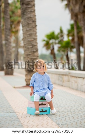 Adorable little girl on a summer vacation sitting on a stack of three small colorful suitcases on a tropical sidewalk with palm trees