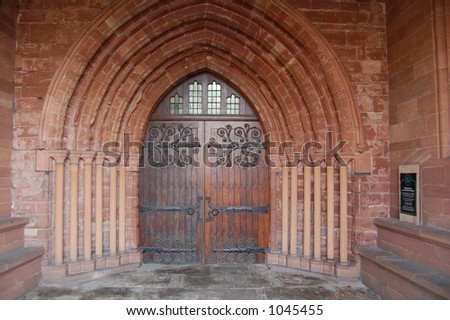 An ancient church door inside a sandstone arch with heavy black iron hinges and a small glass paned panel,with seating inside the porch.