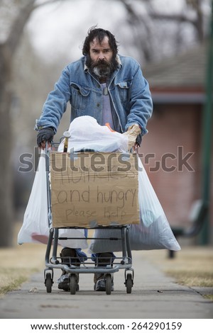 Homeless man with beard pushing a shopping cart with all his possessions.