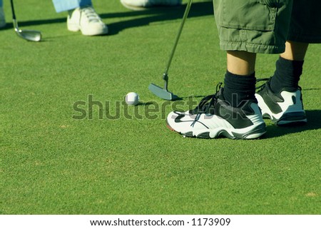 Golf ball on green with putter and feet of player and caddie
