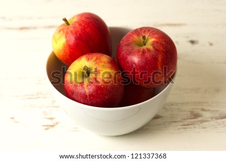 Four red apples in a bowl that's on a white rustic kitchen table.  Apples are healthy and an excellent source of vitamins.  Apples can either be red or green and are crunchy when you bite into them