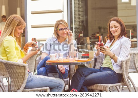 Three Young Women have Coffee Break Together in street cafe. Caucasian female using mobile phone.