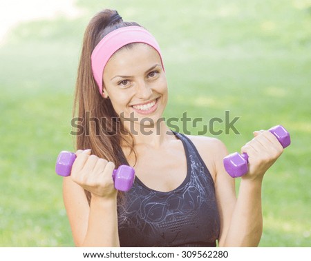 Fitness Slim Woman Training with dumbbells. Smiling attractive female practicing using hand weights outdoor. Healthy lifestyle workout concept on beautiful summer day.