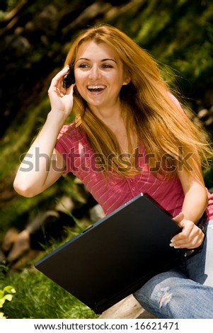 Happy young student using laptop and speaking by phone in natural environment.