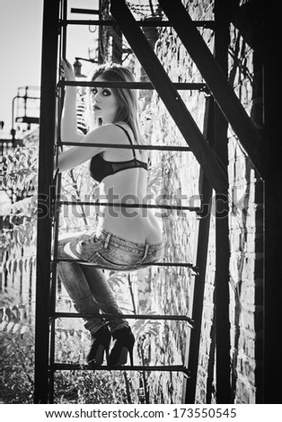 Beautiful young girl in bra and jeans sitting on a metallic staircase. Black and white