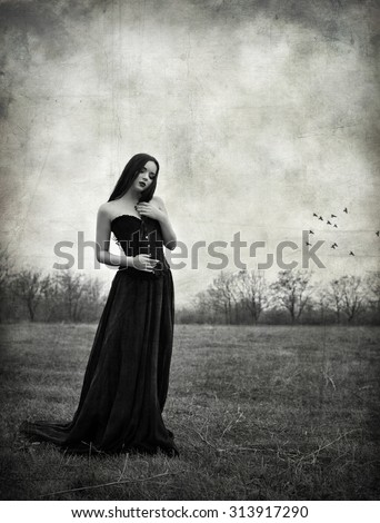 A beautiful sad goth girl stands in autumnal field. Grunge texture effect