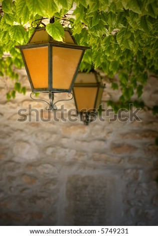 Two black wrought-iron lanterns with orange glass, hanging from a vine-covered ceiling against a stone wall. Taken in an ancient cemetery in France.