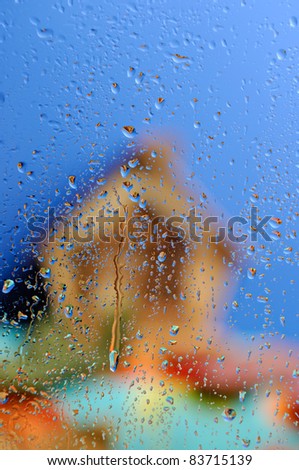 Conceptual stock photo of an Out of focus country house under blue sky behind wet glass in rainy weather