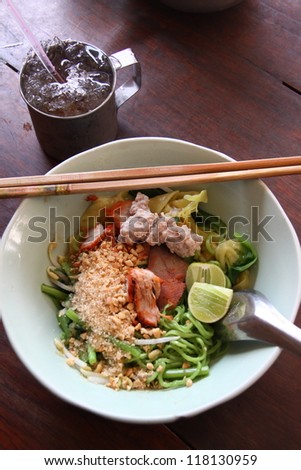 Closeup of asian stir fry with handmade green noodle, pork, vegetables, bean sprouts, and sesame seeds in a white bowl on wooden table.