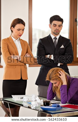 businesswoman holding her head in her hands, sitting at desk, with rumpled paper, coffee, phone, headache and angry colleagues watching her from the back - side view