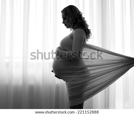 Black and white artistic contre-jour picture of a pregnant woman holding her hands on her belly
