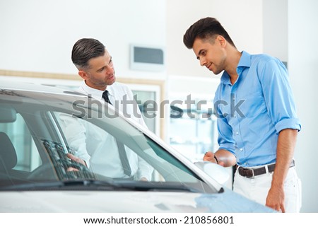 Car Sales Consultant Showing a New Car to a Potential Buyer in Showroom