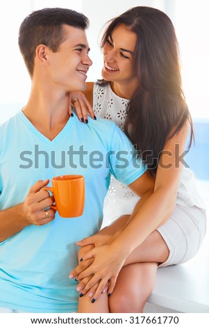 Young happy couple having coffee together at home in the kitchen. Man is holding a cup of coffee and woman leaning on his shoulder.