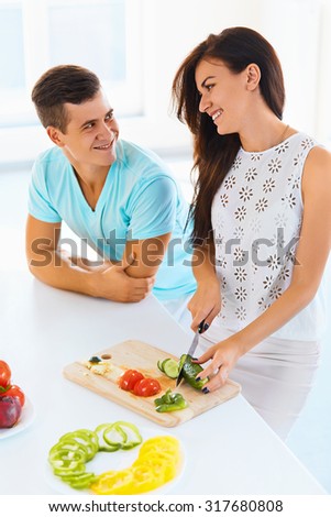 Young couple in love cooking dinner in the kitchen. Woman is cutting vegetables while man looking at her with love.