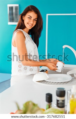 Happy young woman smiling at the camera while washing a cup  in blue kitchen. Housewife enjoying the washing-up