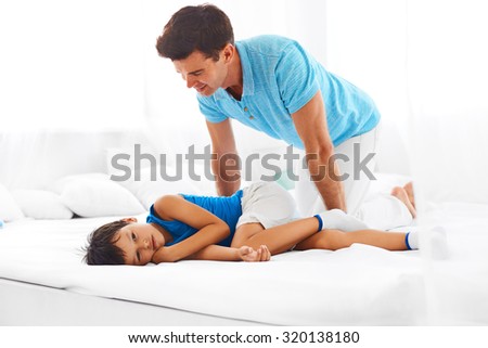 Happy father and sleepy son relaxing on bed together. Boy is resting on the bed after exhausting game and dad is standing over him.