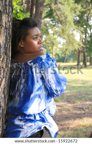 Beautiful African-American female leaning against a tree in a park, summertime setting. Could represent nostalgic, sad feelings, missing someone, thoughtfulness.