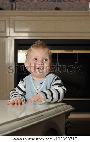 Vertical portrait of a baby boy in a striped sweater and a vest standing next to a coffee table looking up smiling