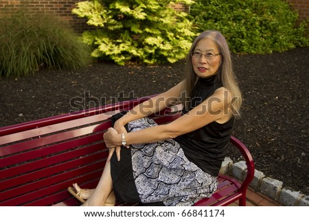 Mature, senior Asian woman seated on a red metal bench has feet up to take a break