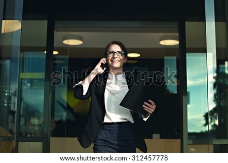Happy successful business woman coming out of the office looking at the sun and smiling. Business people concept. Professional manager talking on smartphone with documents in her hands.