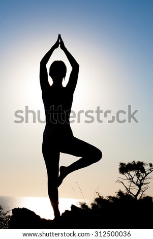 Silhouette of a slender fitness yoga girl in the sun at sunset or sunrise in tree pose. Yoga woman on background of blue sky and blue sea.
